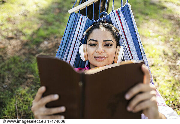 Young woman listening music through headphones reading book in hammock