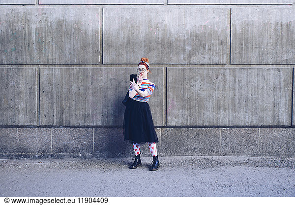 Young woman looking at mobile phone while applying lipstick against wall