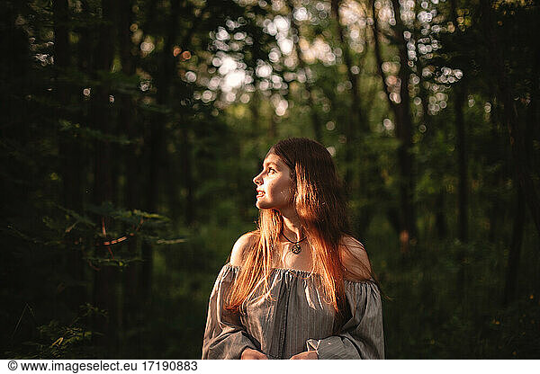 Young woman looking at sunlight while standing in dark forest