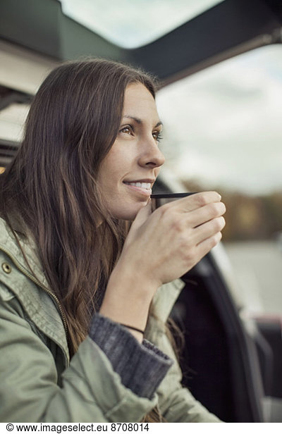 Young woman looking away while drinking coffee at car's trunk