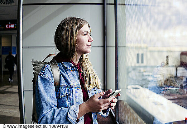 Young woman looking out of window holding smart phone at airport