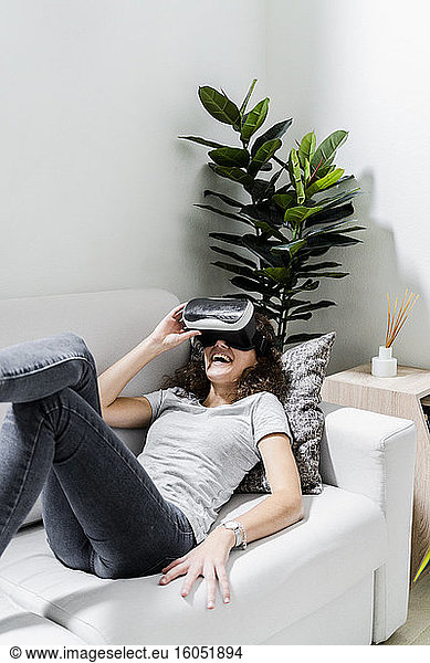 Young woman lying on couch at home using Virtual Reality Glasses