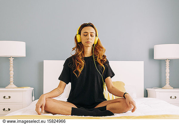 Young woman meditating on bed at home with headphones
