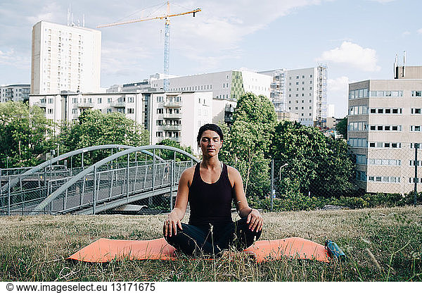 Young woman meditating on field against buildings in city