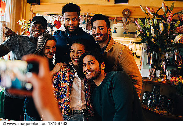 Young woman photographing cheerful multi-ethnic friends standing at restaurant during brunch