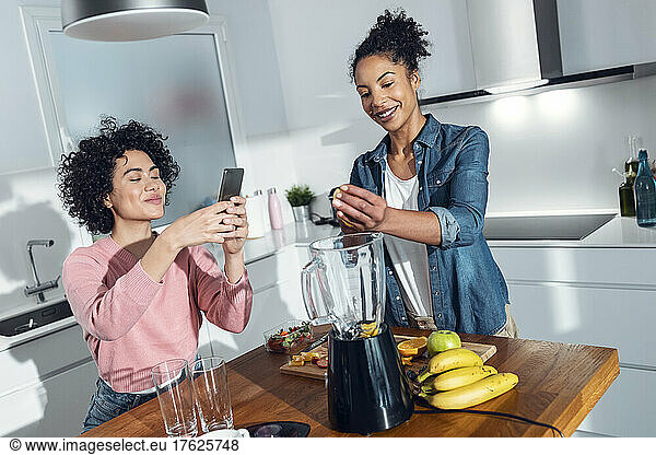 Young woman photographing friend making juice in kitchen at home