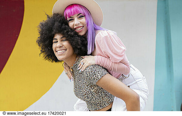 Young woman piggybacking female friend in front of wall