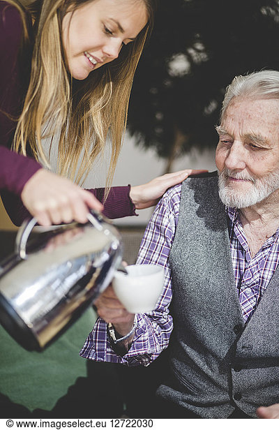 Young woman pouring drink in cup being held by grandfather in nursing home