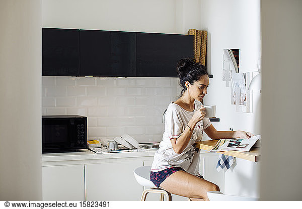 Young woman reading a magazine and drinking coffee in kitchen