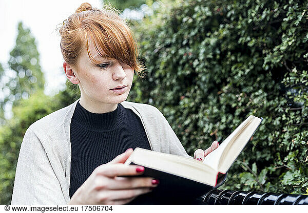 Young woman reading book by hedge