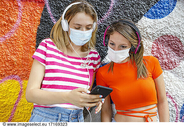 Young woman sharing smart phone with female friend while standing against graffiti wall during pandemic