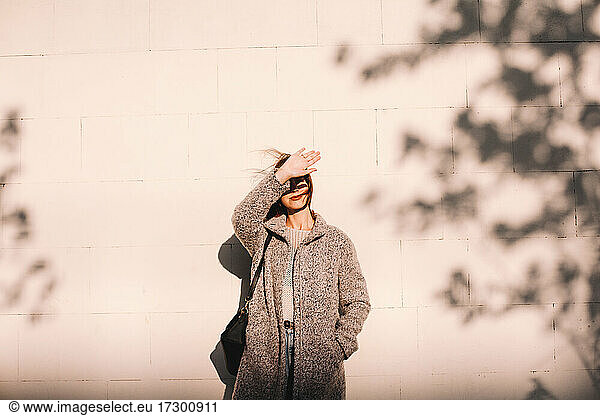 Young woman shielding eyes standing against wall in city