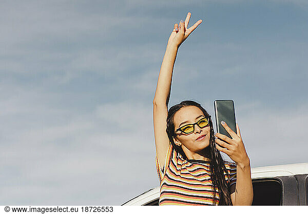 Young woman showing peace sign and taking selfie through smart phone