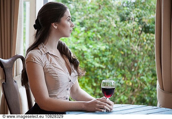 Young woman sitting at table in front of window holding glass of wine looking away