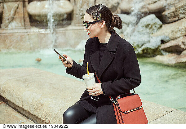 Young woman sitting by the fountain and looking at her phone