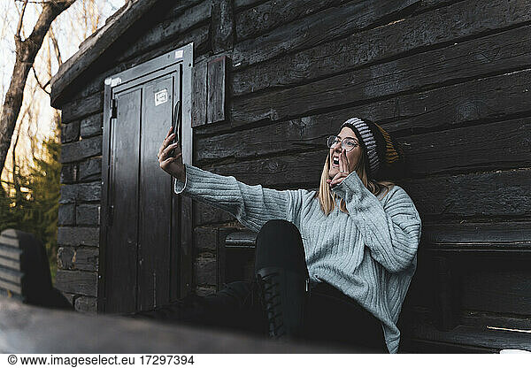 Young woman sitting in front of wooden cabin make a selfie.