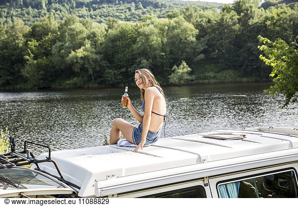 Young woman sitting on roof of a van at lakeside drinking a beer