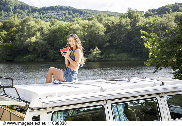 Young woman sitting on roof of a van at lakeside holding slice of watermelon