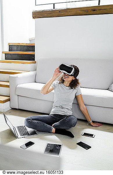 Young woman sitting on the floor at home using Virtual Reality Glasses and various electronic devices