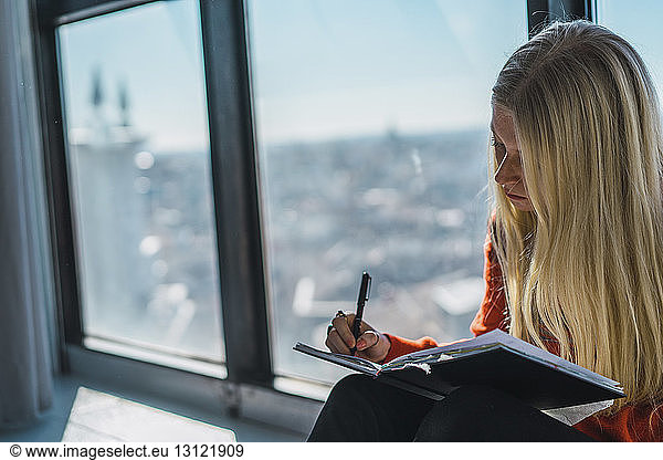 Young woman sketching on book while sitting against windows at home
