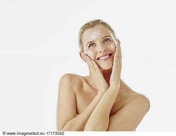 Young woman smiling while applying face cream standing against white background