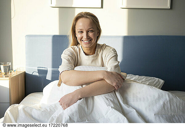 Young woman smiling while hugging knees on bed at home