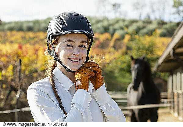 Young woman smiling while wearing helmet in front of horse at paddock