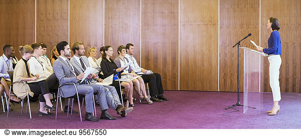 Young woman standing at transparent lectern  talking before audience in conference room