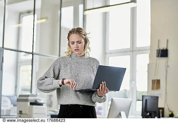 Young woman standing in office with laptop checking the time