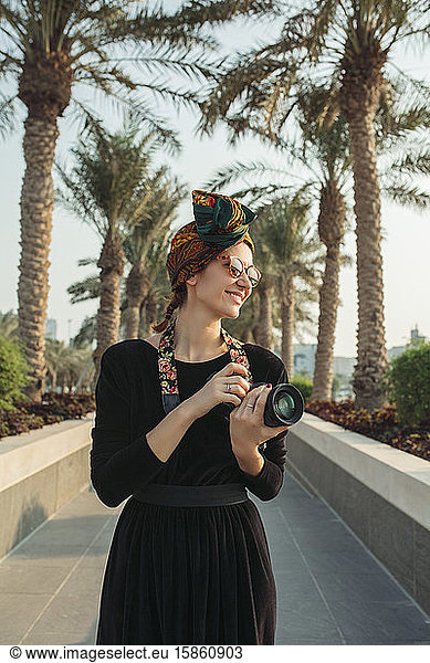 Young woman standing in palm alley wearing headwrap and holding camera