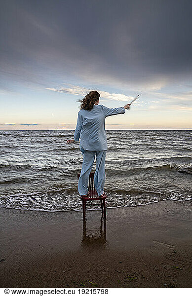 Young woman standing on chair and gesturing with stick in front of sea
