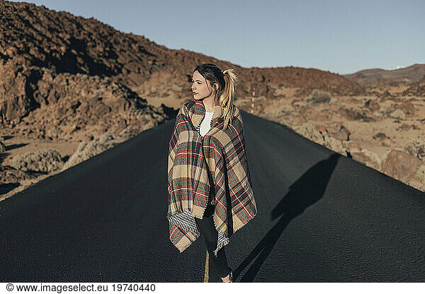 Young woman standing on road near volcanic landscape wrapped in blanket