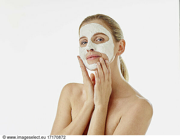 Young woman staring while applying face mask standing against white background