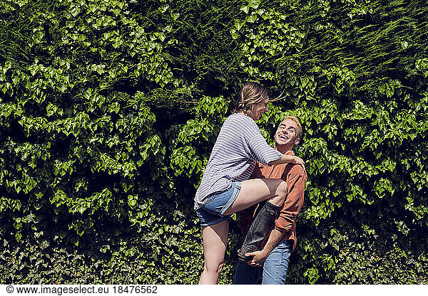 Young woman stepping on boyfriend's hands in front of hedge
