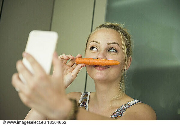 Young woman taking a selfie and making moustache with a carrot on her face  Munich  Bavaria  Germany