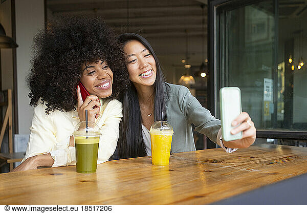 Young woman taking selfie with friend talking on smart phone at cafe