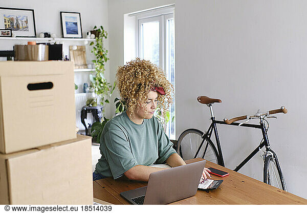 Young woman using calculator by laptop at desk