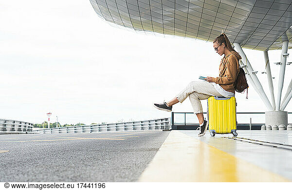Young woman using digital tablet while sitting on wheeled luggage at airport
