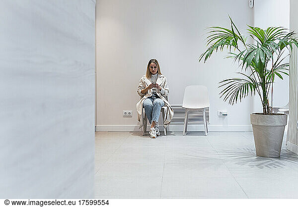 Young woman using mobile phone sitting in waiting room at clinic