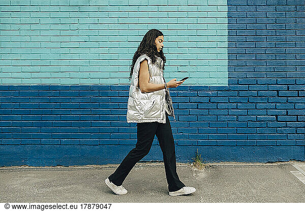Young woman using mobile phone walking by blue brick wall