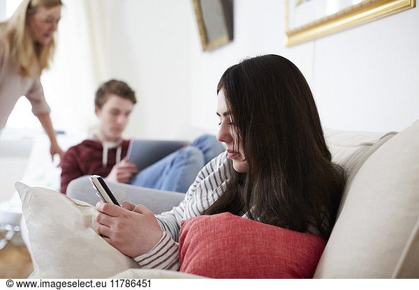 Young woman using mobile phone while mother looking at son with laptop in living room