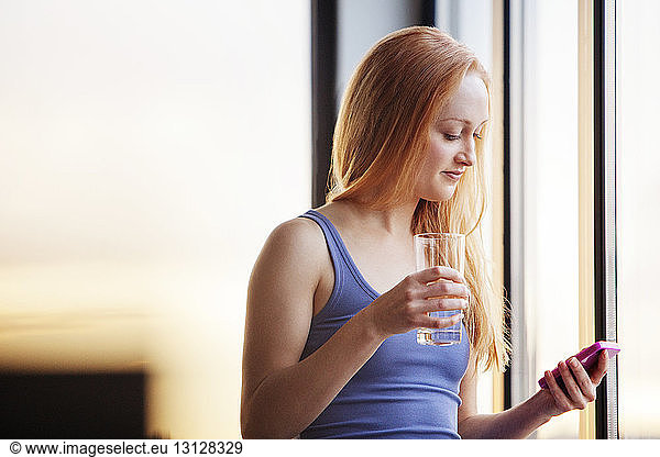 Young woman using phone while holding drinking glass at home