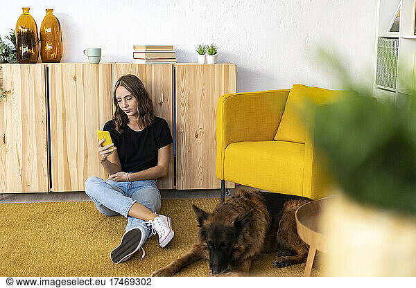 Young woman using smart phone by German Shepherd in living room