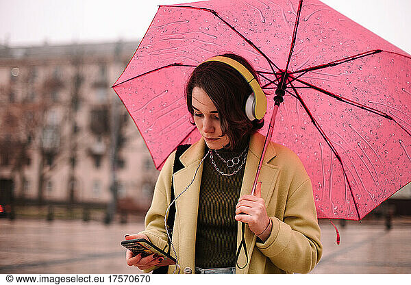 Young woman using smart phone holding umbrella standing in city