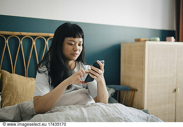 Young woman using smart phone on bed in bedroom