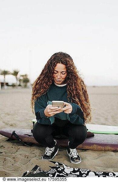 Young woman using smart phone sitting on surfboard at beach