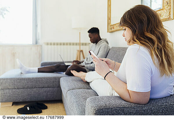 Young woman using smart phone sitting with boyfriend on sofa at home