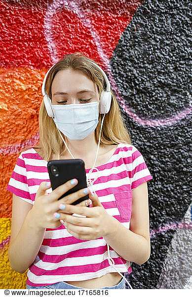 Young woman using smart phone while listening music through headphones against graffiti wall during pandemic