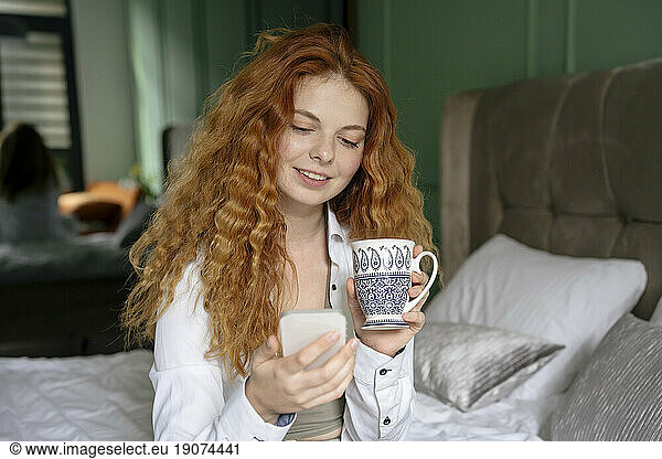 Young woman using smart phone with tea cup sitting on bed
