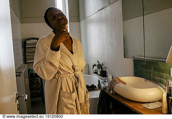 Young woman wearing bathrobe using jade roller on face in bathroom